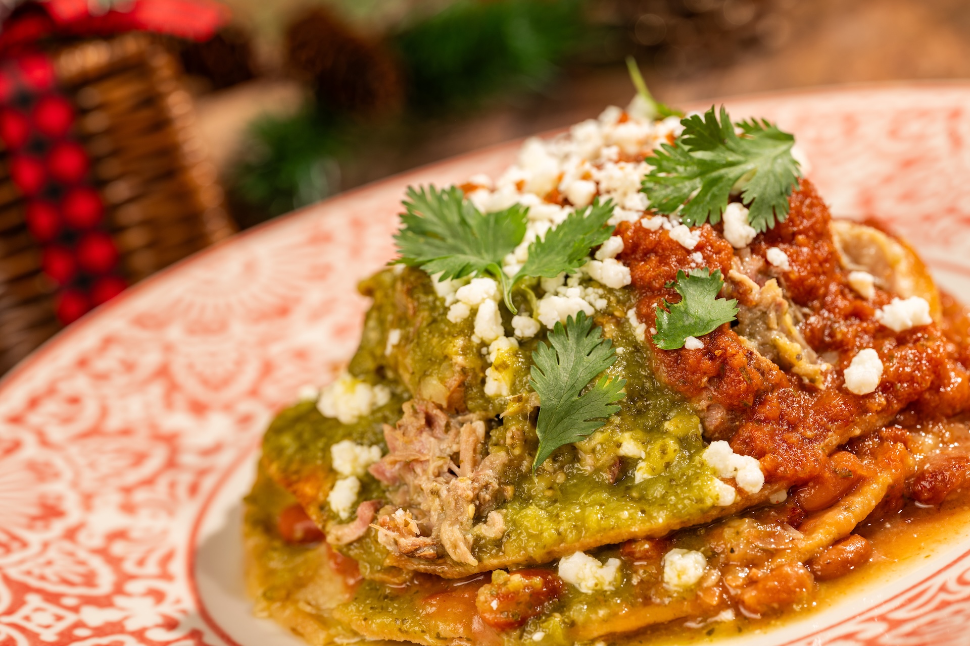 A tostada piled with shredded smoked pork, green salsa verde and red tomato salsa, topped with crumbled white queso fresco and cilantro leaves, on a red and white patterned plate