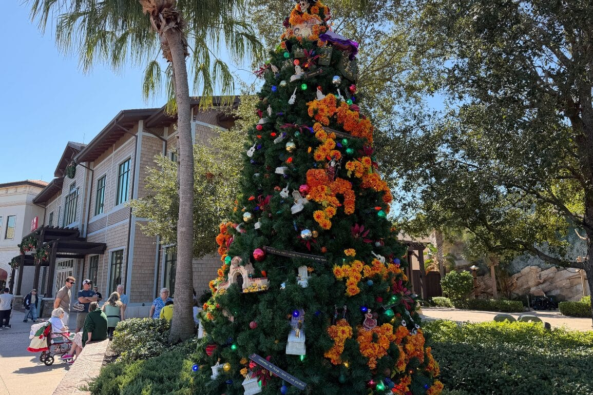 A Christmas tree covered in orange marigold garlands, small skeleton figures, guitars and colored lights, standing beside a palm tree and a shingled Disney Springs building