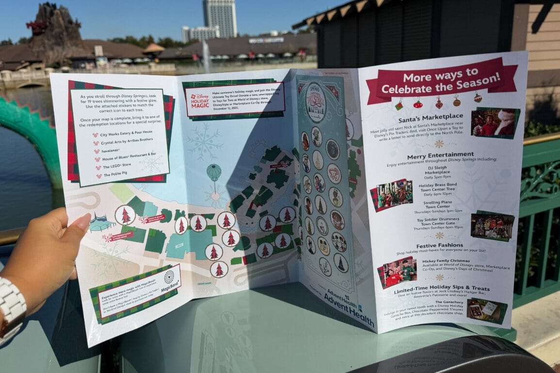 A hand holding open the 2025 Christmas Tree Stroll map, showing tree icons on a Disney Springs map, a column of sticker spots and a More ways to Celebrate the Season panel, with the Disney Springs waterfront behind it