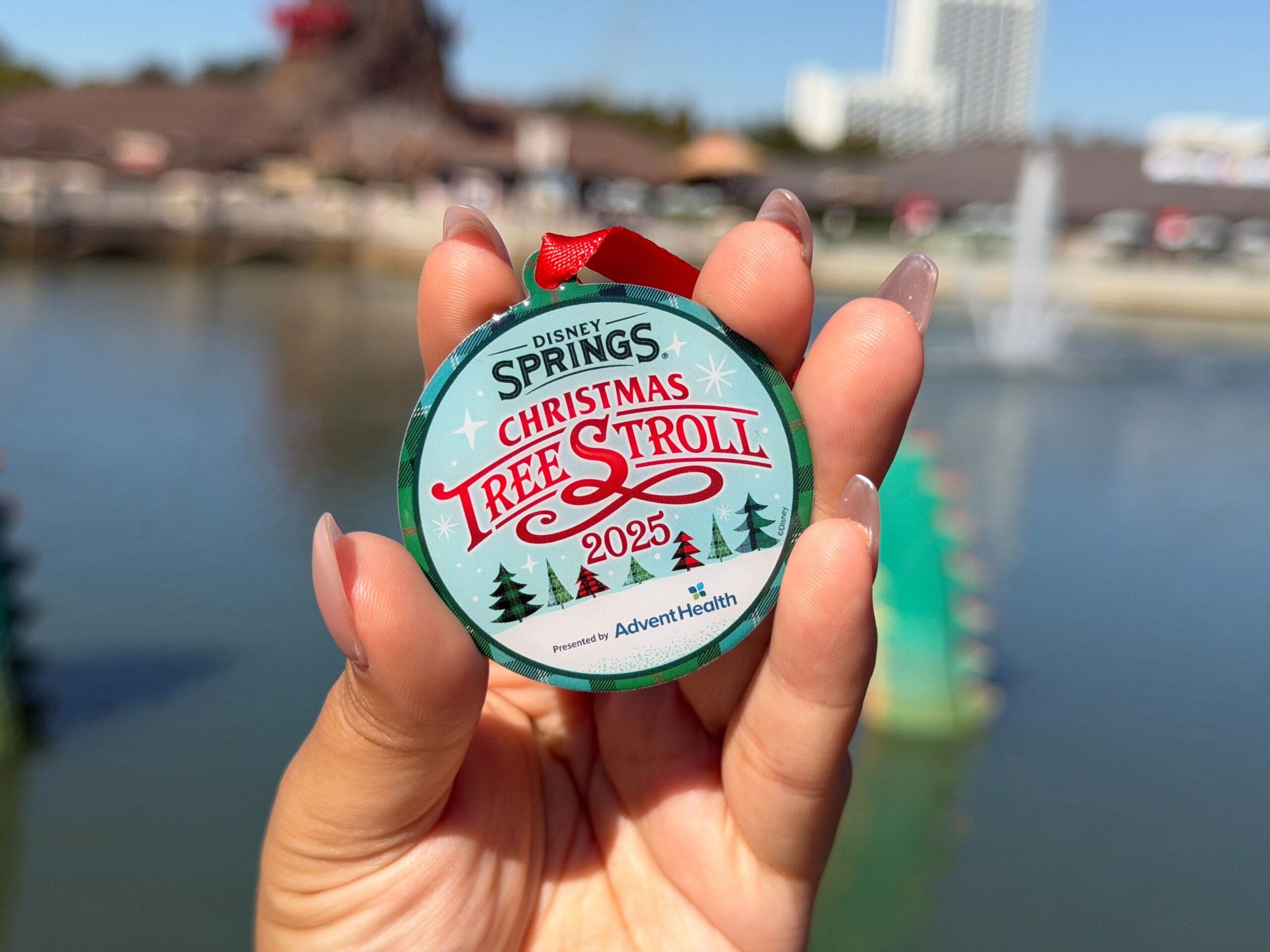 A hand holding a round green and blue plaid ornament on a red ribbon loop, its face reading Disney Springs Christmas Tree Stroll 2025, presented by AdventHealth, with the Disney Springs waterfront blurred behind it