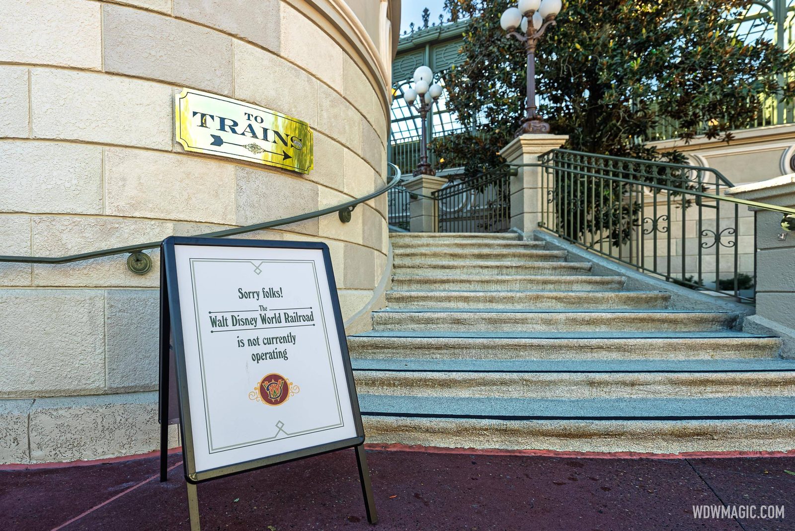 A sandwich board reading Sorry folks! The Walt Disney World Railroad is not currently operating, at the foot of the stone staircase to the Main Street train station, beside a brass To Trains sign on the curved wall