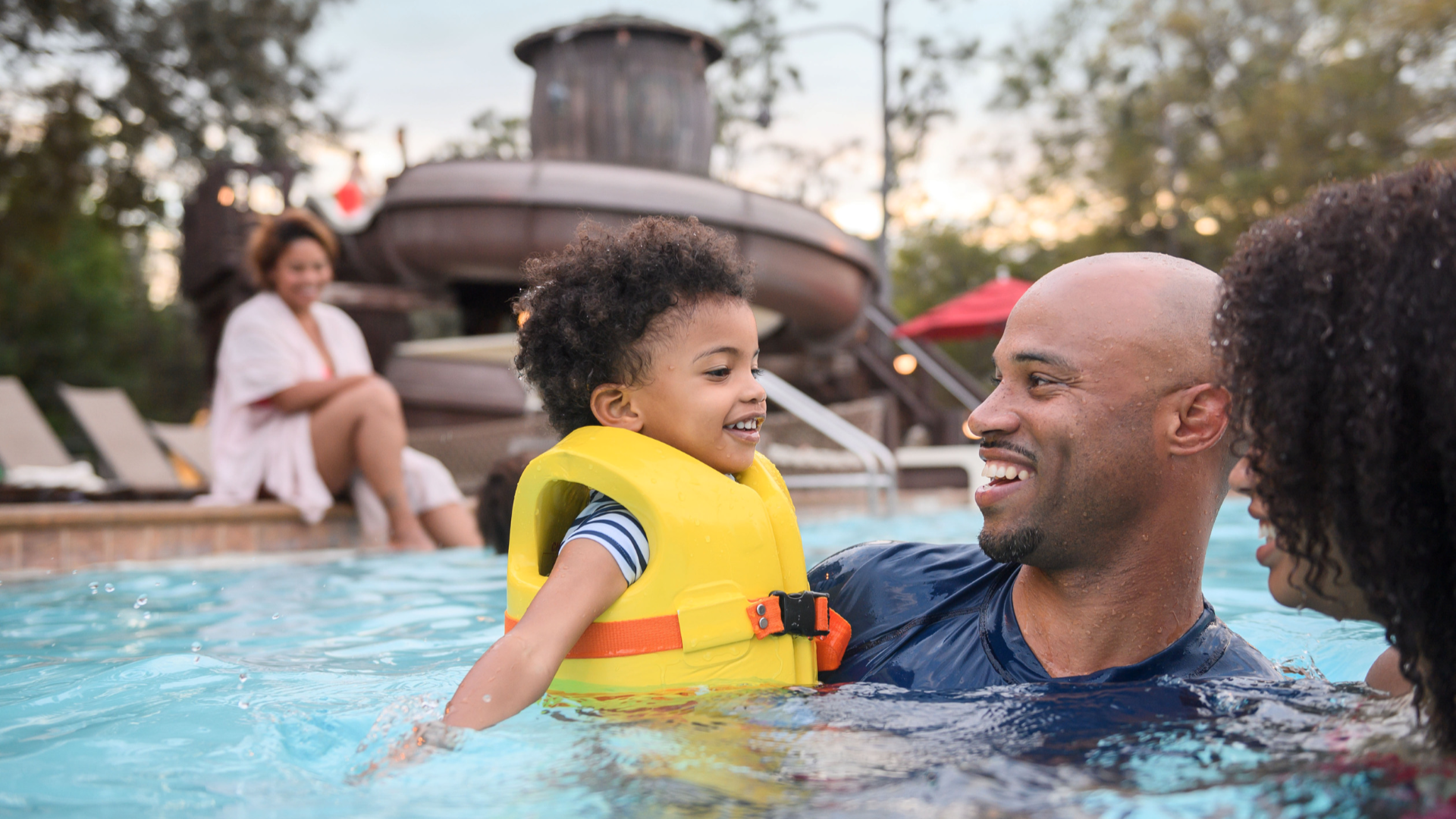A father and small child in a life vest playing in the pool at Fort Wilderness, with the wooden waterslide tower visible behind them