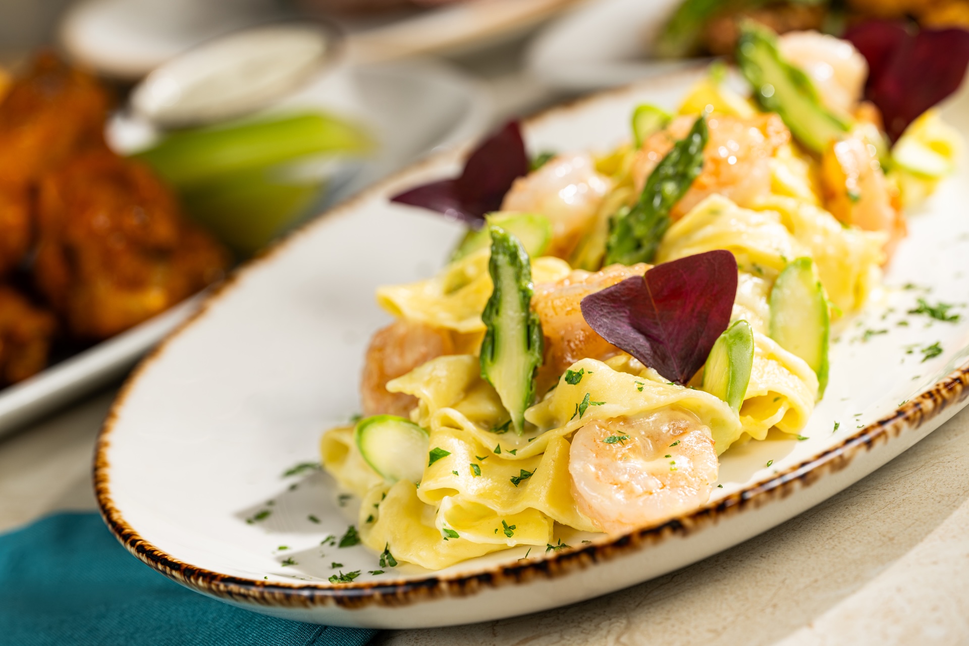 Pappardelle pasta with shrimp, asparagus and purple leaf garnish on an oval plate, with chicken wings behind it