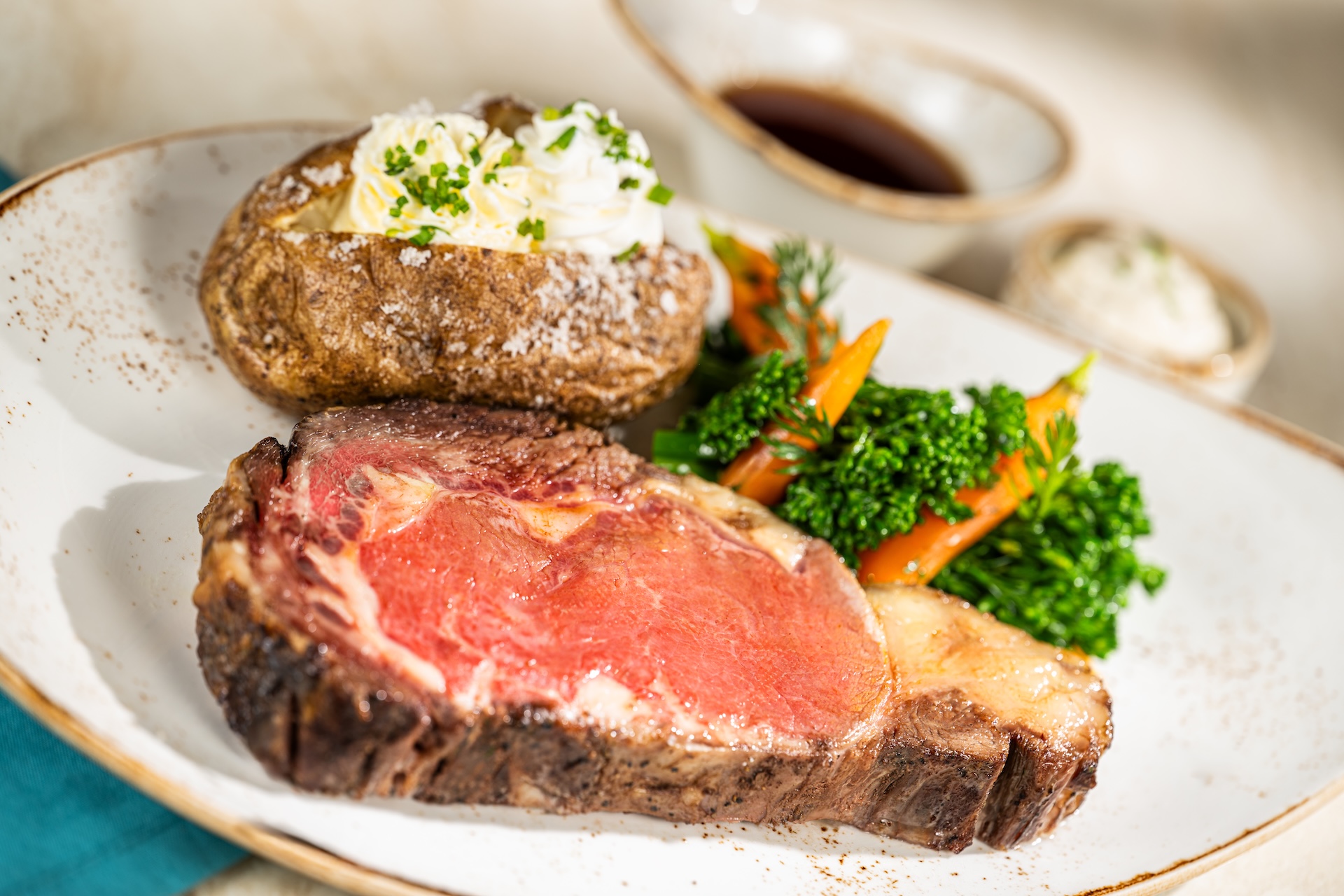 Slow-roasted prime rib with a baked potato and seasonal vegetables, with two sauce ramekins behind the plate